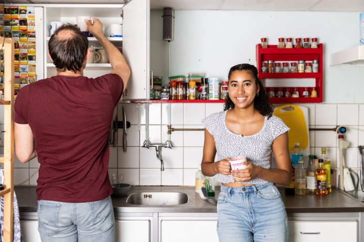Twee studenten staan in de keuken van hun studentenhuis. Een drinkt een kop koffie, de ander zoekt iets in een kastje.