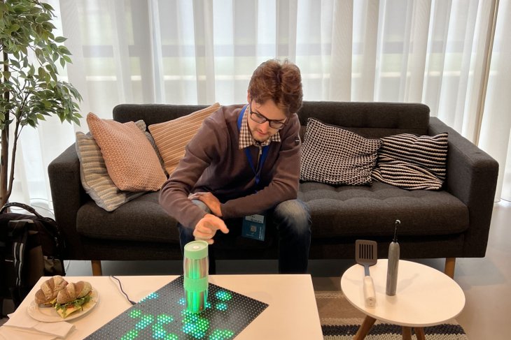 Een man zit op een bank en experimenteert met een apparaat op een tafel dat groene LED-lichten activeert. Op de tafel liggen ook een sandwich en diverse kleine objecten, waaronder een elektrische tandenborstel en een keukengerei.