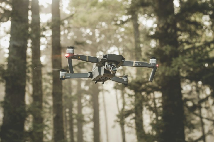 A drone hovering in mid-air among tall pine trees in a misty forest, with soft sunlight filtering through the trees.