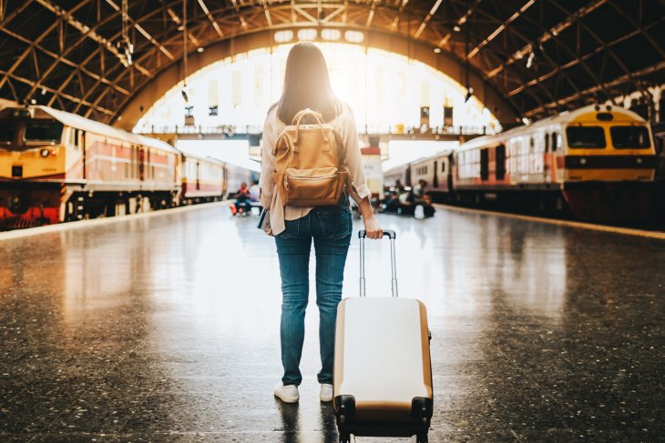 A student standing at the trainstation with a suitcase in hand.