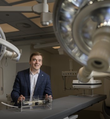 Een glimlachende man in pak staat in een moderne operatiekamer en houdt een transparant prototype van een medisch apparaat vast op een operatietafel, met grote operatielampen en medische apparatuur op de achtergrond.