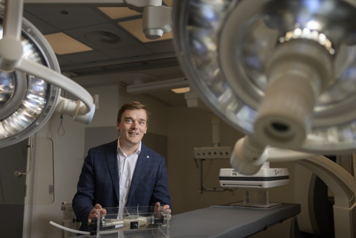 A smiling man in a suit stands in a modern operating room, holding a transparent medical device prototype on a surgical table, with large surgical lamps and medical equipment in the background.