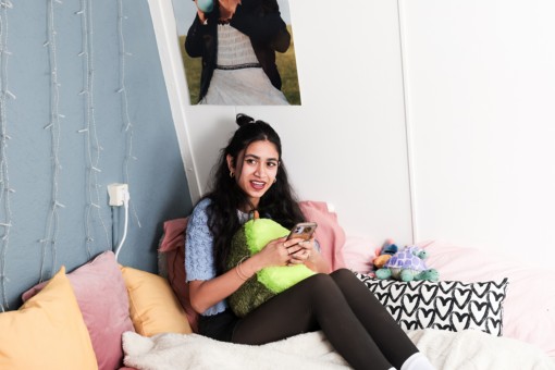 A student sitting on her bed in her room. She has fairy lights and posters up the wall.