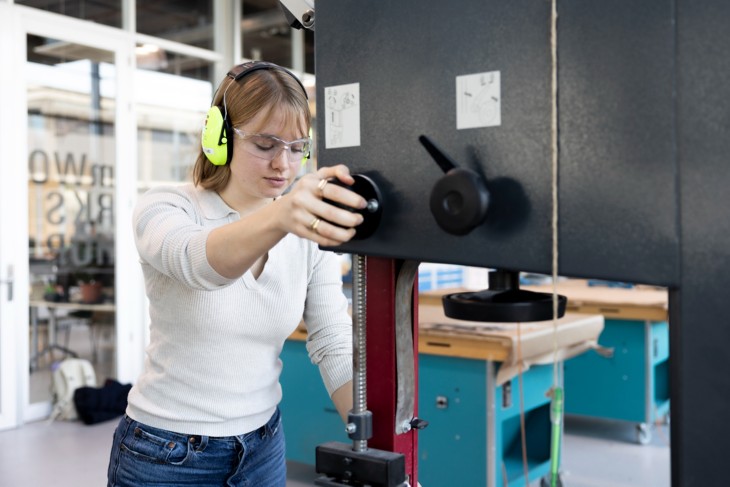 Girl in a workshop, wearing safety goggles and ear protection, operating a machine.
