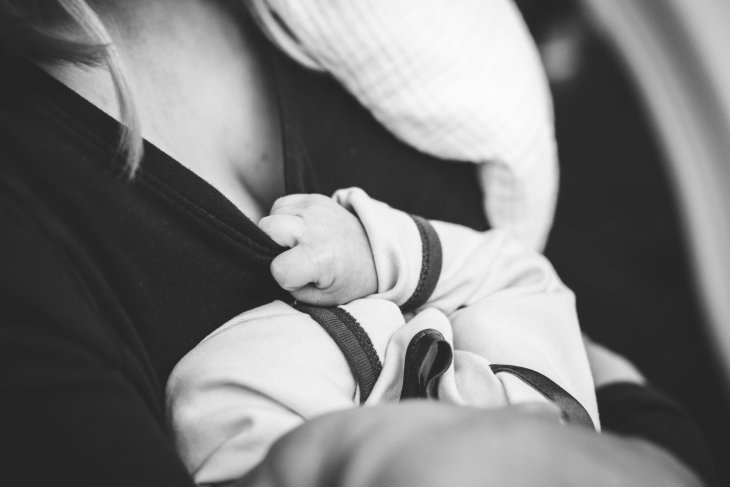 Black and white close-up of a baby breastfeeding, gently gripping the mother's clothing. The scene captures an intimate and loving moment.
