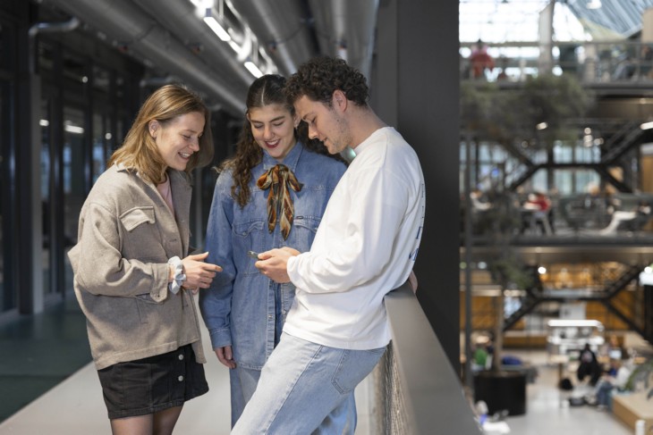 Three students laugh together, looking at a phone.