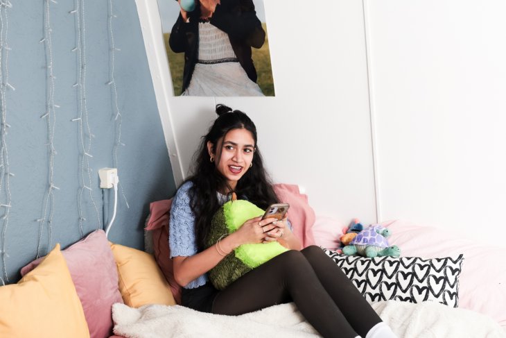 Een student zit op haar bed in haar kamer. Ze heeft kerstverlichting en posters aan de muur hangen.
