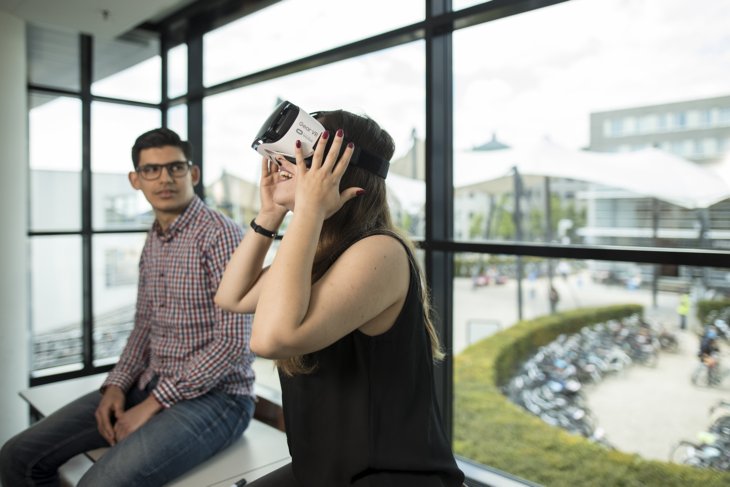 A student is sitting near a window watching a fellow student use VR glasses