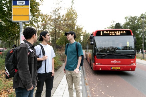 A group of three students waits at the bus stop. A bus drives up to them.