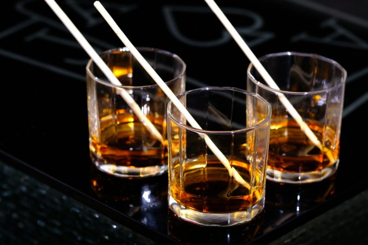 Three glasses filled with amber-coloured whisky, each with a straw, placed on a glossy black table with blurred white symbols in the background.