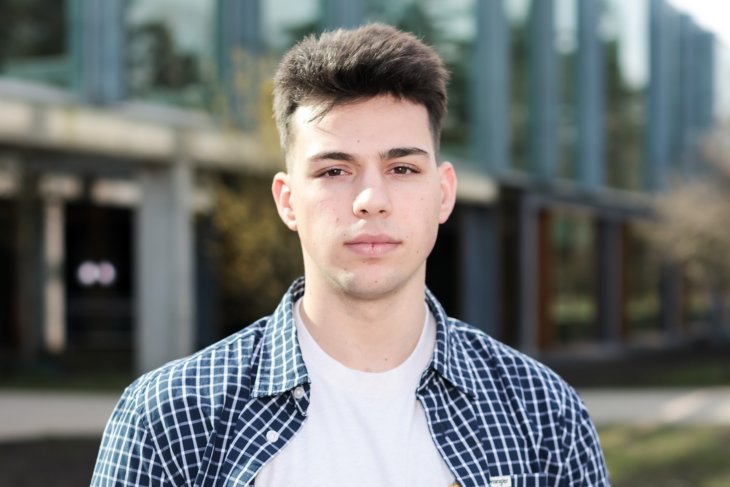 Marios voor het Langezijds gebouw op de campus, kijkend in de camera.