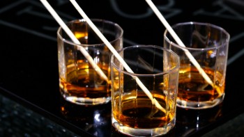 Three glasses filled with amber-coloured whisky, each with a straw, placed on a glossy black table with blurred white symbols in the background.
