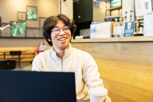 Student Wisang sitting behind his laptop and smiling to someone behind the camera.