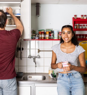 Twee studenten staan in de keuken van hun studentenhuis. Een drinkt een kop koffie, de ander zoekt iets in een kastje.
